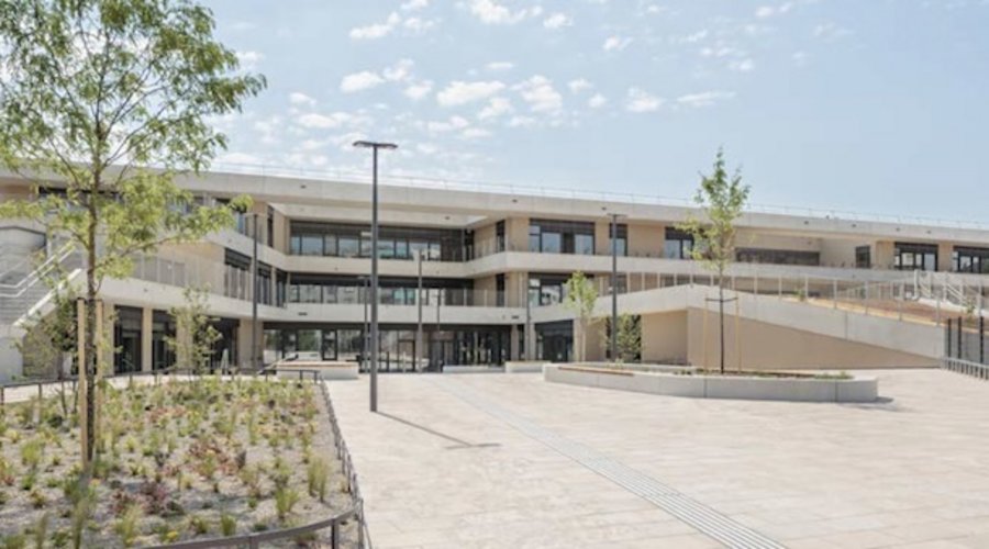 Moderne mehrstöckige Betonfassade mit Glasfronten, umgeben von gepflasterten Flächen und jungen Bäumen unter blauem Himmel.