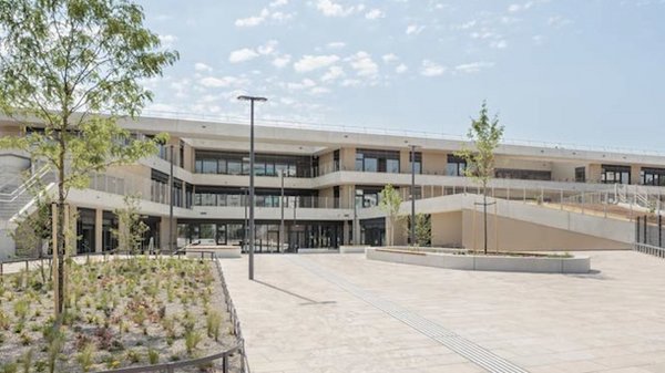 Moderne mehrstöckige Betonfassade mit Glasfronten, umgeben von gepflasterten Flächen und jungen Bäumen unter blauem Himmel.